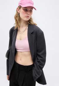 Νεαρή γυναίκα που φοράει ροζ καπέλο, ροζ κοντό μπλουζάκι, σκουρόχρωμο φαρδύ σακάκι, μαύρο παντελόνι, ασημένιες αλυσίδες και κρατά μαύρη τσάντα ώμου, κοιτάζοντας προς τα κάτω αριστερά.