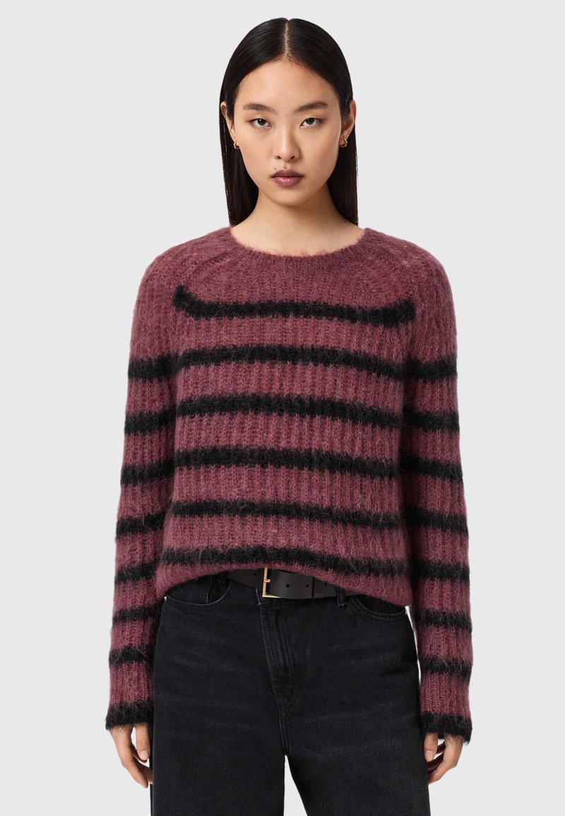 Femme portant un pull flou mauve avec des rayures horizontales noires et un pantalon noir, debout devant un fond uni gris clair.