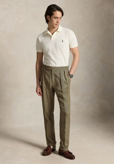 Mężczyzna ubrany w białą koszulkę polo z krótkim rękawem, brązowe spodnie w kratę z wysokim stanem, brązowe mokasyny z frędzlami oraz zegarek na rękę, stojący na neutralnym tle.