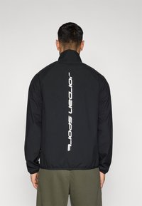 Μαύρο αδιάβροχο μπουφάν με ψηλό γιακά, με κάθετο λευκό λογότυπο "Jordan Sport" στην πλάτη και ελαστικές μανσέτες στα μανίκια.