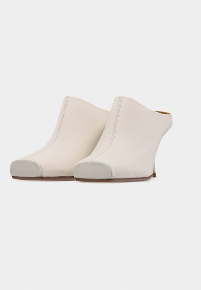 Ζευγάρι λευκές δερμάτινες μπότες mule με στρογγυλεμένες μύτες από σουέτ και τακούνια μπλοκ σε απλό ανοιχτό φόντο.