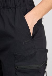 Des pantalons cargo noirs légers avec une taille élastique, une poche latérale et une poche zippée avec un logo de marque discret. Tissu texturé.