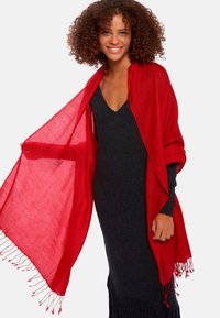 Écharpe rouge en tissu léger, posée sur les épaules avec des bords frangés. Portée sur une robe sombre et ajustée, affichant une texture lisse.