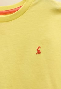 T-shirt jaune à col rond avec petit logo de lapin rouge brodé sur le côté gauche de la poitrine et un bord intérieur de l'encolure orange.