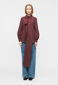 Blouse bordeaux rayée avec un devant asymétrique, un col boutonné et des manches longues bouffantes, associée à un pantalon large en denim bleu.