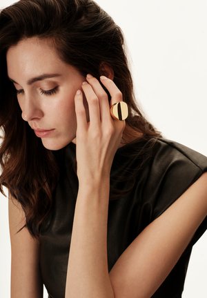 Vrouw met donker haar die een zwart mouwloos topje draagt, haar gezicht aanraakt en een grote ronde gouden ring aan haar vinger toont.