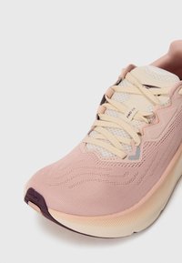 Baskets de sport rose clair avec une tige en maille texturée, des lacets crème, une semelle rembourrée et une semelle extérieure violet foncé. Présente un branding subtil.