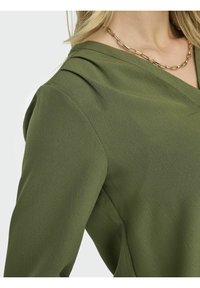 Olive green top with a v-neckline and textured fabric. Features soft draping at the shoulder and a gold chain necklace accent.