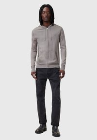 Felpa zip grigia chiara con tasca frontale, abbinata a jeans scuri e stivaletti neri. Presenta un cappuccio con coulisse e un dettaglio logo minimale.