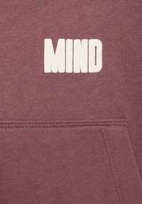 Primo piano di una felpa bordeaux con la parola "MIND" in lettere bianche in grassetto, con dettagli sulla tasca frontale e una trama di tessuto morbido.