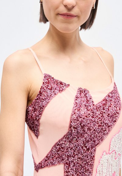 Γυναίκα που φοράει ένα ροζ αμάνικο τοπ με λαμπερά ροζ και κόκκινα παγιέτα και λεπτούς ιμάντες στους ώμους, μπροστά σε λευκό φόντο.