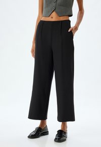 Zwarte wijde broek met plooien, gemaakt van een soepel materiaal. Ze hebben zijzakken en een subtiele glans, gecombineerd met glanzende zwarte schoenen.