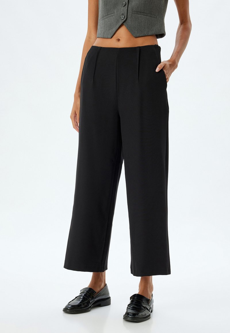 Zwarte wijde broek met plooien, gemaakt van een soepel materiaal. Ze hebben zijzakken en een subtiele glans, gecombineerd met glanzende zwarte schoenen.