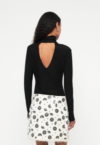 Žena s kovrčavom kosom nosi crnu majicu dugih rukava s velikim izrezom na leđima i bijelu suknju s crnim cvjetnim uzorkom.