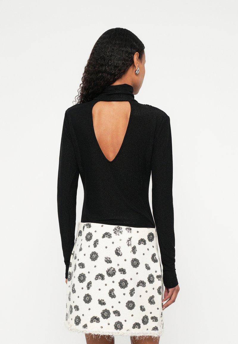 Žena s kovrčavom kosom nosi crnu majicu dugih rukava s velikim izrezom na leđima i bijelu suknju s crnim cvjetnim uzorkom.