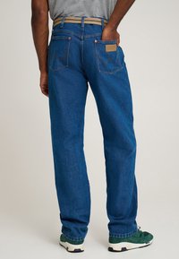 Blugi din denim albastru cu croială dreaptă, buzunare posterioare vizibile și o etichetă de piele cu brand. Purtați cu adidași verzi și albi.