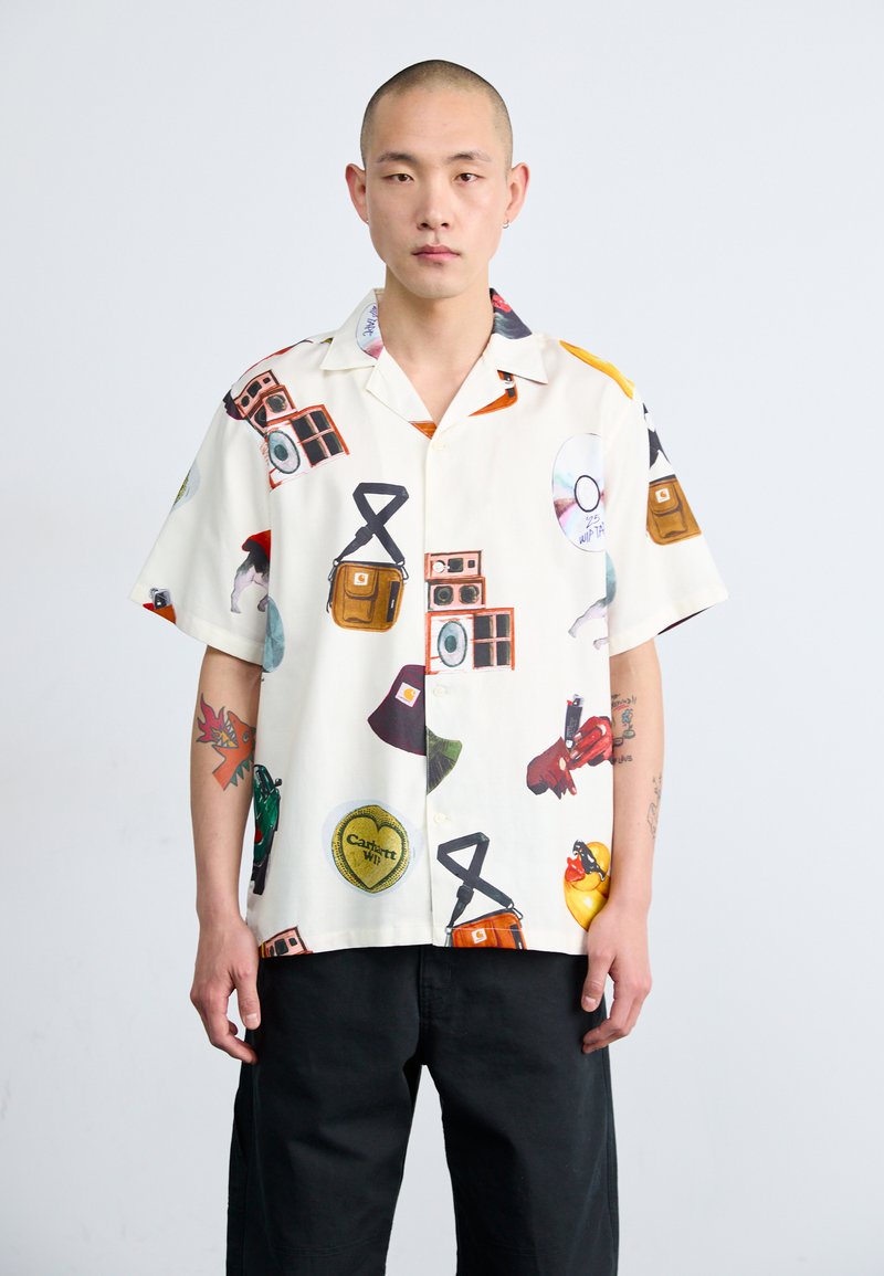 Camisa de manga corta color crema con diversos gráficos estampados, incluyendo cámaras y bolsas. Fabricada con tejido ligero y de corte relajado.