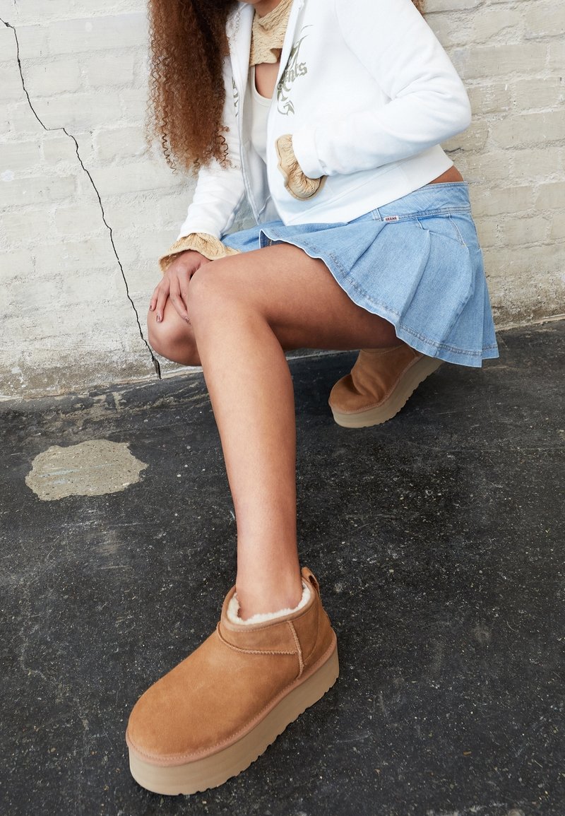 Jeune personne accroupie contre un mur de briques blanches fissuré, portant une veste blanche, une jupe en denim bleu et des chaussures compensées couleur beige doublées en shearling.
