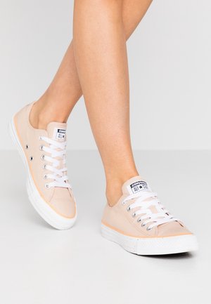Světle růžové nízké tenisky Converse s bílými tkaničkami, nošené s překříženýma nohama na jednobarevném bílém pozadí.