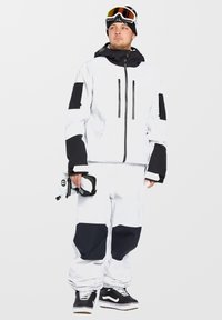 Hvid og sort skijakke med hætte og lynlås-lommer, parret med matchende bukser med sorte accenter. Snowboardbriller holdt i den ene hånd.
