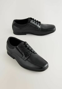 Dos pares de zapatos de vestir de cuero negro con cordones, exhibidos desde arriba sobre un fondo claro. Los zapatos tienen un diseño elegante y pulido.