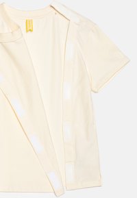 Světle žluté tričko vyrobené z jemné bavlny, s kulatým výstřihem a bočním uzavřením na suchý zip pro snadné oblékání. Hladká textura a jednoduchý design.