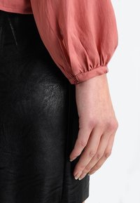 Blusa rosa con maniche a sbuffo e polsini arricciati, abbinata a una gonna in pelle nera. Texture liscia e sottile lucentezza visibile su entrambi i capi.
