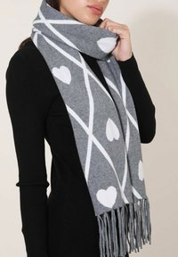 Écharpe grise avec des motifs de cœurs blancs et des rayures diagonales, dotée de bords à franges et d'une texture douce. Portée autour du cou.