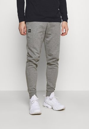 Homme portant un pantalon de survêtement gris Under Armour et des baskets blanches, debout devant un fond blanc uni.
