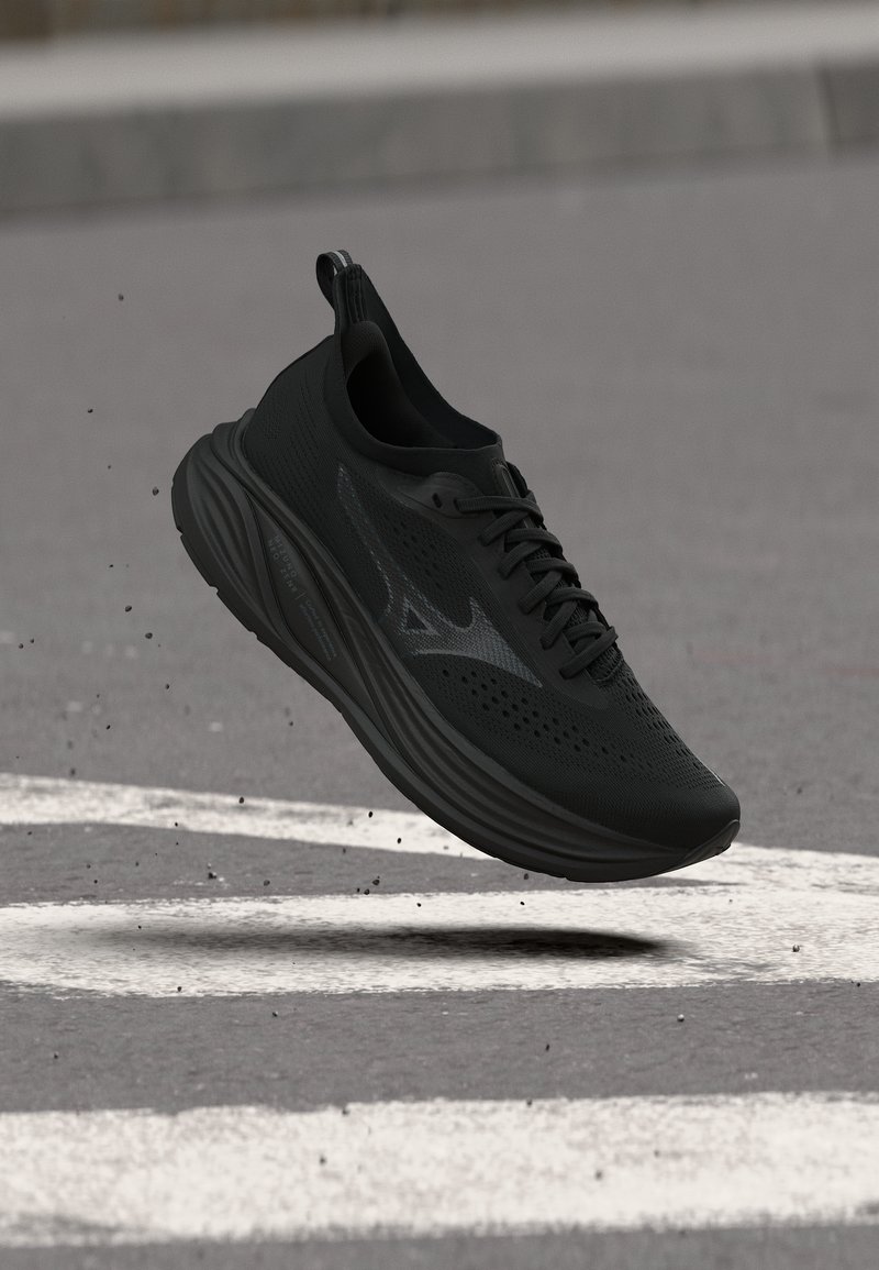 Μαύρο αθλητικό παπούτσι τρεξίματος με παχιά σόλα που αιωρείται πάνω από έναν ασφαλτοστρωμένο δρόμο με λευκές οδικές σημάνσεις.