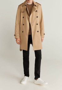 Beige Trenchcoat mit dopp Reihen-Design, Knopfakzenten und Schulterklappen, getragen über einem braunen Pullover und schwarzen Hosen, kombiniert mit weißen Sneakers.