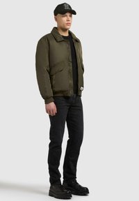 Olivgrüne Bomberjacke mit Reißverschluss, zwei Fronttaschen, schwarzes T-Shirt, dunkle Jeans und schwarze Schnürstiefel mit dicken Sohlen.