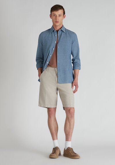 Camisa azul claro de botones con mangas remangadas, pantalones cortos beiges, zapatillas marrones y calcetines blancos, exhibidos sobre un fondo neutro.