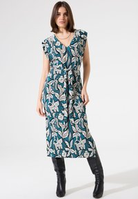 Maxikleid mit floralem Muster in Teal, featuring einen V-Ausschnitt, Rüschensäumen und einer gebundenen Taille. Getragen mit schwarzen knielangen Stiefeln.