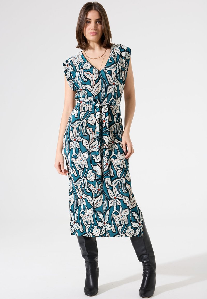 Maxikleid mit floralem Muster in Teal, featuring einen V-Ausschnitt, Rüschensäumen und einer gebundenen Taille. Getragen mit schwarzen knielangen Stiefeln.