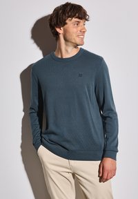 Mörk teal crew-neck tröja i mjukt tyg, med långa ärmar och en liten logotyp på bröstet. Matchas med ljusa byxor.