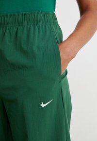 Hand rustend in de zak van donkere groene Nike shorts met elastische tailleband en wit Nike swoosh logo op de dij.