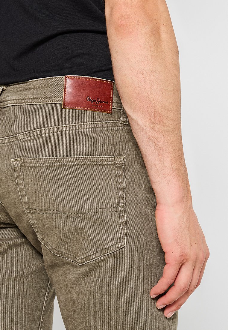 Pantalones caquis de corte slim, con bolsillos traseros y una etiqueta de marca de cuero marrón cosida en la cintura. Textura suave.