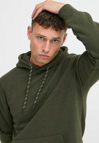 Hoodie vert olive avec capuche à cordon, poche avant et poignets côtelés. Le matériau semble doux avec une texture lisse.