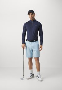 Marine langermet skjorte med glidelåskrage, lyseblå shorts og svart belte. Modellen holder en golfkølle og har på seg svart caps og sko.