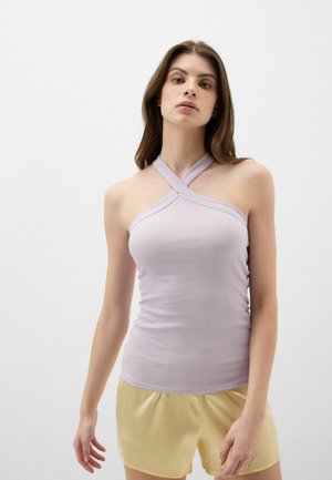 Ung kvinde iført en lyselilla ribbet halternecktop med krydsede stropper og lysegule shorts, stående mod en ensfarvet hvid baggrund.