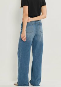 Jeans en denim bleu à jambes larges avec un délavage, cinq poches et ourlets effilochés, associés à une chemise noire. Vue arrière du pantalon.