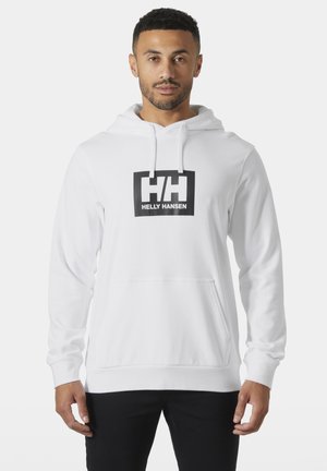 Hombre con una sudadera blanca Helly Hansen con bolsillo frontal y pantalones negros, de pie contra un fondo blanco liso.