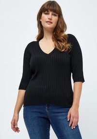 Schwarzer Rippenpullover mit V-Ausschnitt und kurzen Ärmeln. Kombiniert mit blauen Jeans, die eine figurbetonte Silhouette und ein strukturiertes Material zeigen.