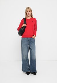 Felpa rossa in cotone con scollatura rotonda e emblema a forma di cuore, abbinata a jeans in denim blu a gamba larga e una borsa a tracolla nera. Texture liscia.