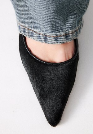 Zapato de tacón negro con punta afilada, hecho de material texturizado y con un borde brillante, mostrando parte de un pie y unos vaqueros de mezclilla deshilachados en el fondo.