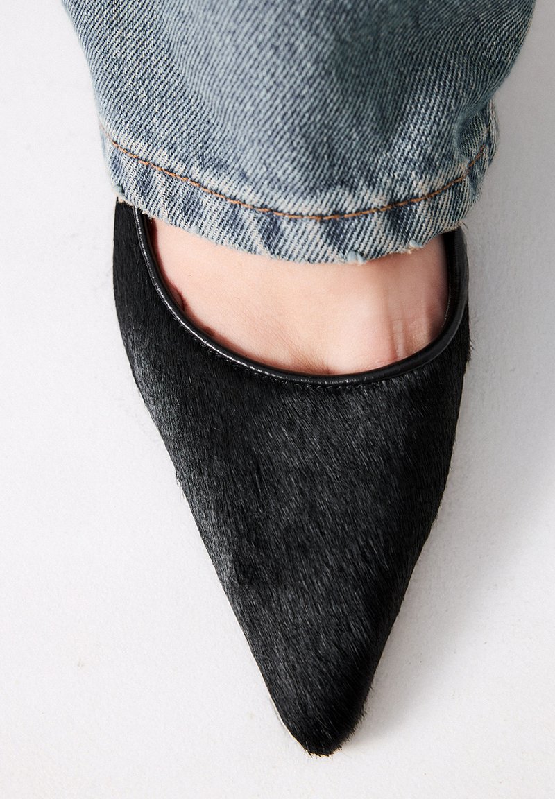 Zapato de tacón negro con punta afilada, hecho de material texturizado y con un borde brillante, mostrando parte de un pie y unos vaqueros de mezclilla deshilachados en el fondo.