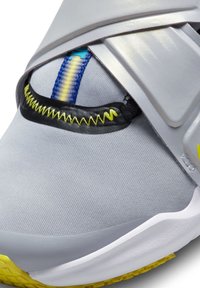 Chaussure de sport avec une tige synthétique grise, une bande élastique, des accents jaunes et une semelle texturée pour une bonne adhérence. Détails de coutures bleues et jaunes.