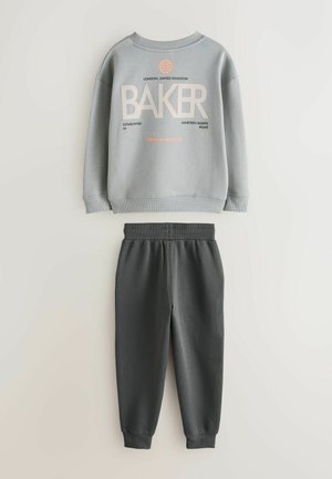 Grijze trui met de tekst "BAKER" en grafische accenten, gecombineerd met donkergrijze joggingbroek met elastische tailleband en taps toelopende boorden.