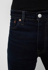 Mörkblå denimjeans med en figurnära design. Funktioner inkluderar fem fickor, kopparfärgade nitar och kontrasterande sömmar längs sömmarna.